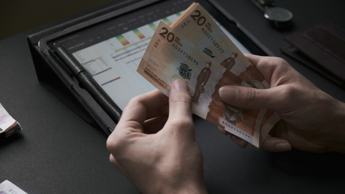 Dos manos contando billetes de 20 mil pesos colombianos con una tablet en el fondo.